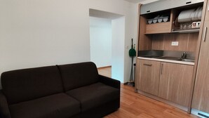 A modern living room with a dark sofa, wooden flooring, a kitchen with wooden cabinets, and a ceiling light fixture. - B&B Berninapass (Poschiavo)