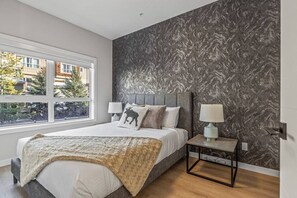 A bedroom with a bed, two bedside tables with lamps, a window with a view of buildings and trees, and a textured wall. - 2BR Townhome with Mountain View 3min from DT Canmore (Canmore)
