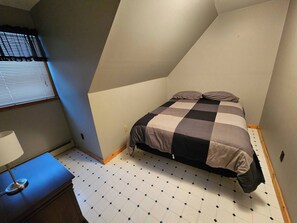 Ein Schlafzimmer mit kariertem Bettbezug, einem Nachttisch mit Lampe und einem Fenster mit Jalousien.