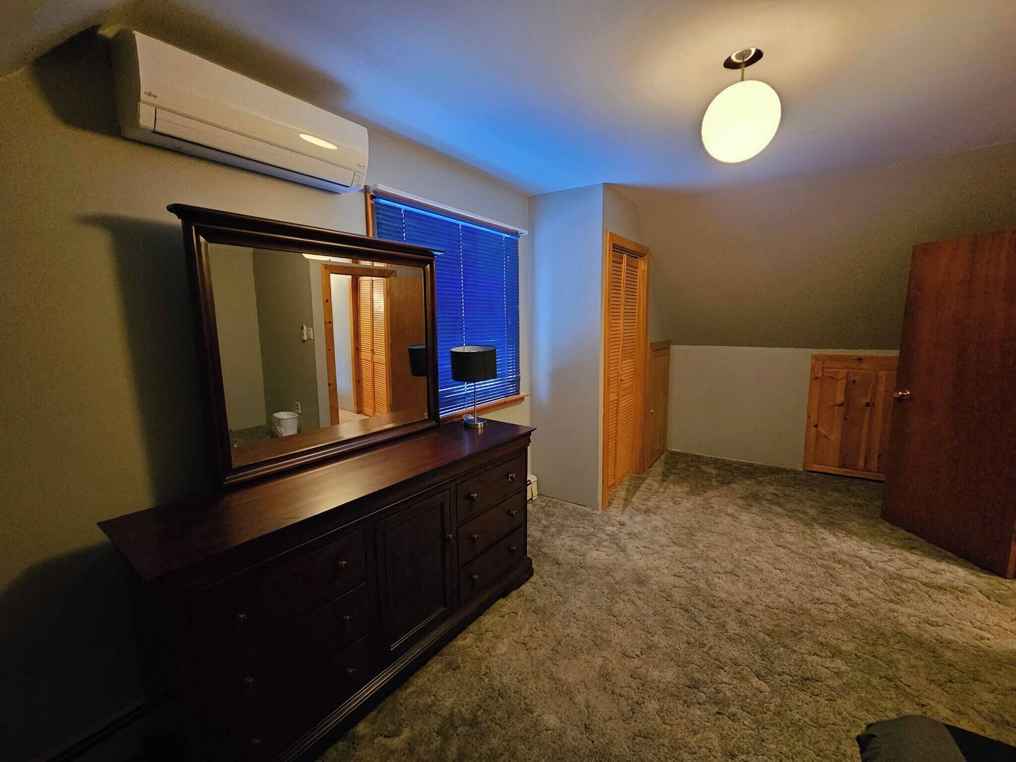 Een slaapkamer met een kapstok, een spiegel, een plafondlamp, een raam met zonwering, een houten deur en een vloerbedekking.