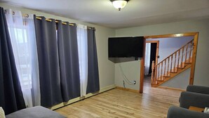 A living room with a flat-screen TV mounted on the wall, a wooden staircase, and a grey sofa. - Bayview Hideaway is a 2 Story Home With Lots of Privacy (Whycocomagh)