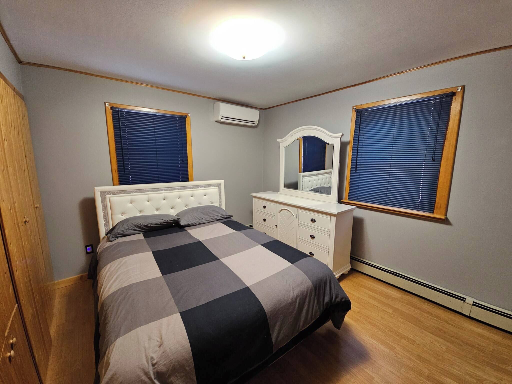 Une chambre à coucher comprenant un lit, une commode, un miroir et deux fenêtres équipées de stores.