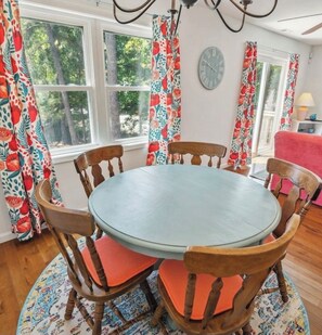 A round dining table with six wooden chairs, each with a red cushion. The room has large windows with floral curtains, a clock on the wall, and a chandelier.