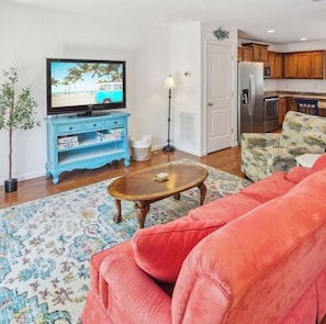 A living room with a television, a blue entertainment unit, a red sofa, a patterned rug, and a kitchen in the background.