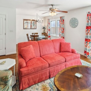 A living room with a red sofa, a wooden coffee table, a ceiling fan, a dining area with wooden chairs, and a window with floral curtains.