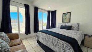 Ein Schlafzimmer mit Bett, Sessel, Nachttisch, Fenster mit Vorhängen und einem Bild an der Wand.