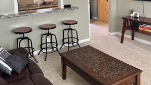 Un salon avec un canapé en cuir marron, une table basse, trois tabourets de bar, un ventilateur de plafond, un téléviseur et un coin cuisine doté de meubles en bois et d’une fenêtre.