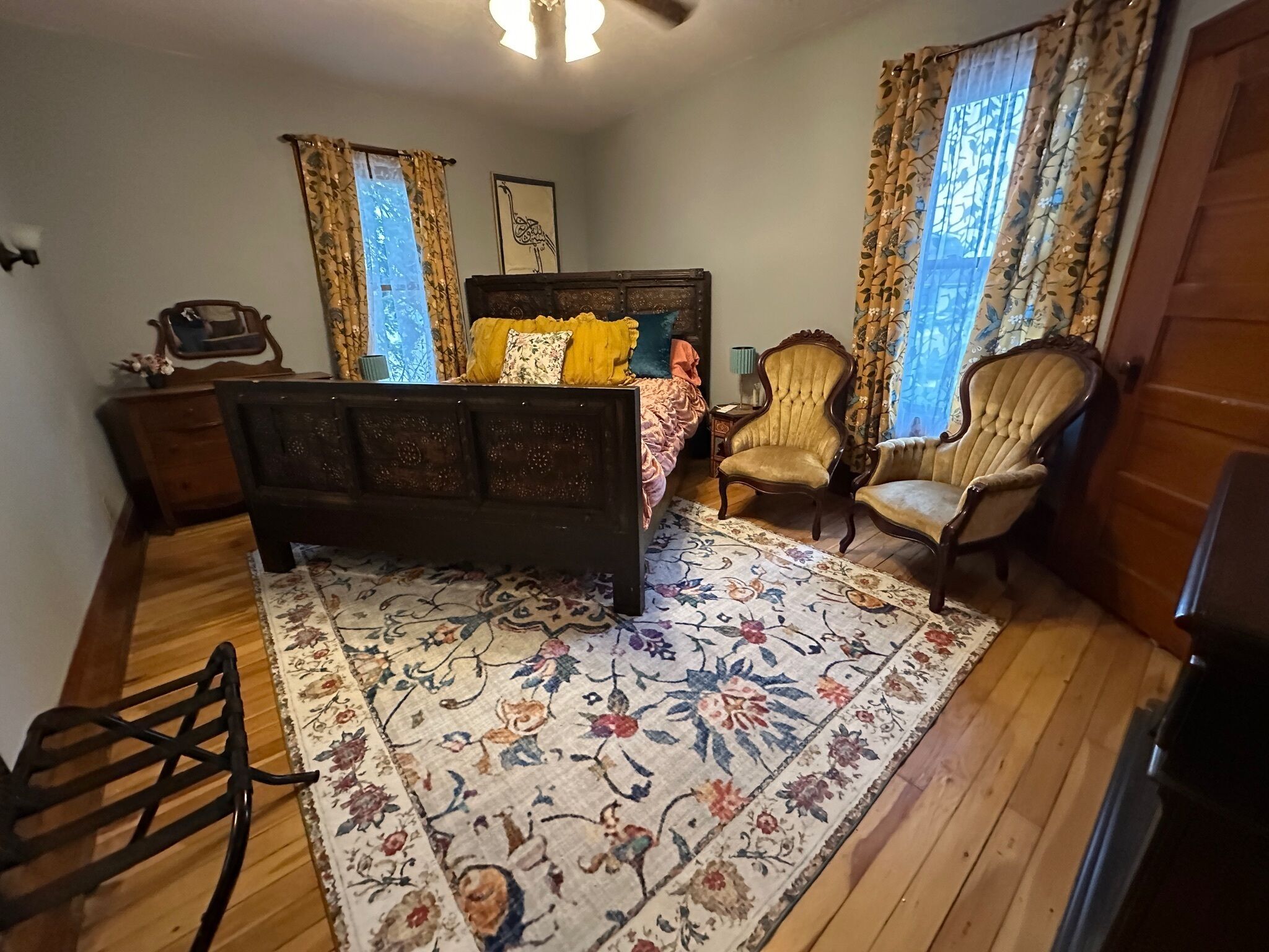 A bedroom with a bed, two armchairs, a wooden dresser, a nightstand, and a floral rug.