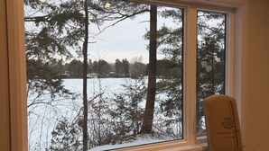 A room with a large window overlooking a snowy landscape, a dark sofa, and a wooden chair. - Cozy waterfront 2-bedroom cottage in peaceful Seeley's Bay with AC (Seeley's Bay)