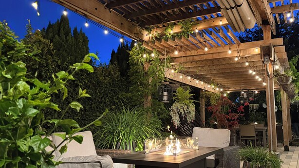 Um pátio externo com pérgola de madeira, luzes de cordão, móveis de vime e lareira.