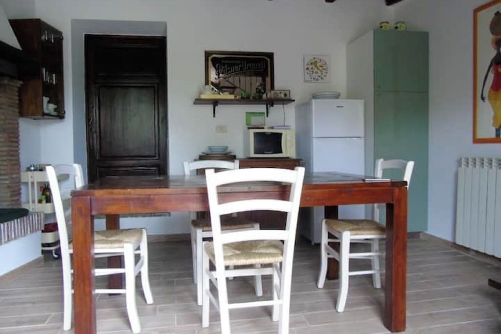 Comedor con mesa de madera y cuatro sillas, microondas, refrigerador y un estante con diversos objetos.