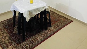 Un coin repas avec une table recouverte d’une nappe blanche, un tapis à motifs et un tableau représentant un paysage accroché au mur.