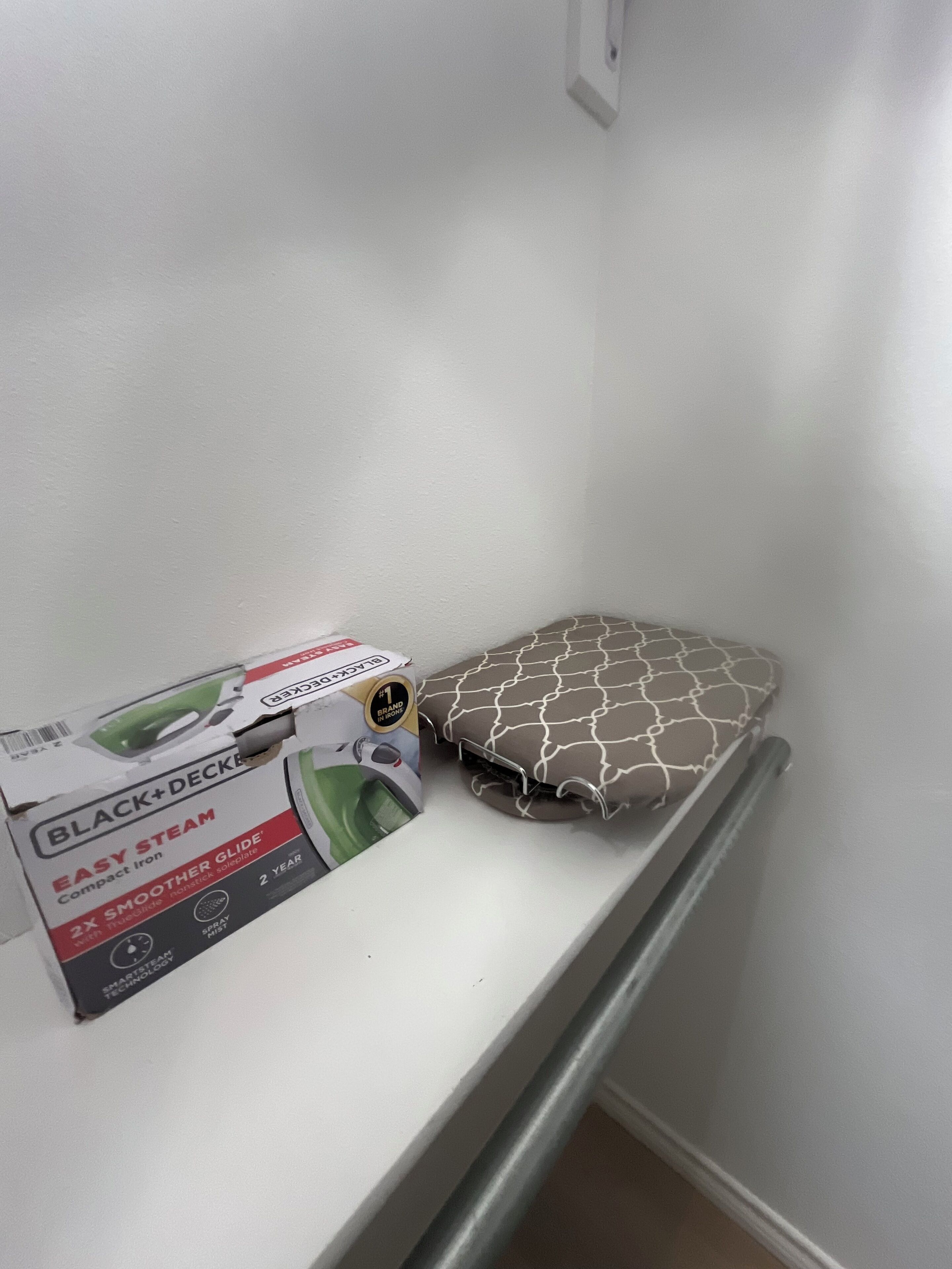 A corner of a room with a white wall, a metal shelf, and a box labeled 'BLACK+DECKER' with a '2X SMOOTHER GLIDE' iron.