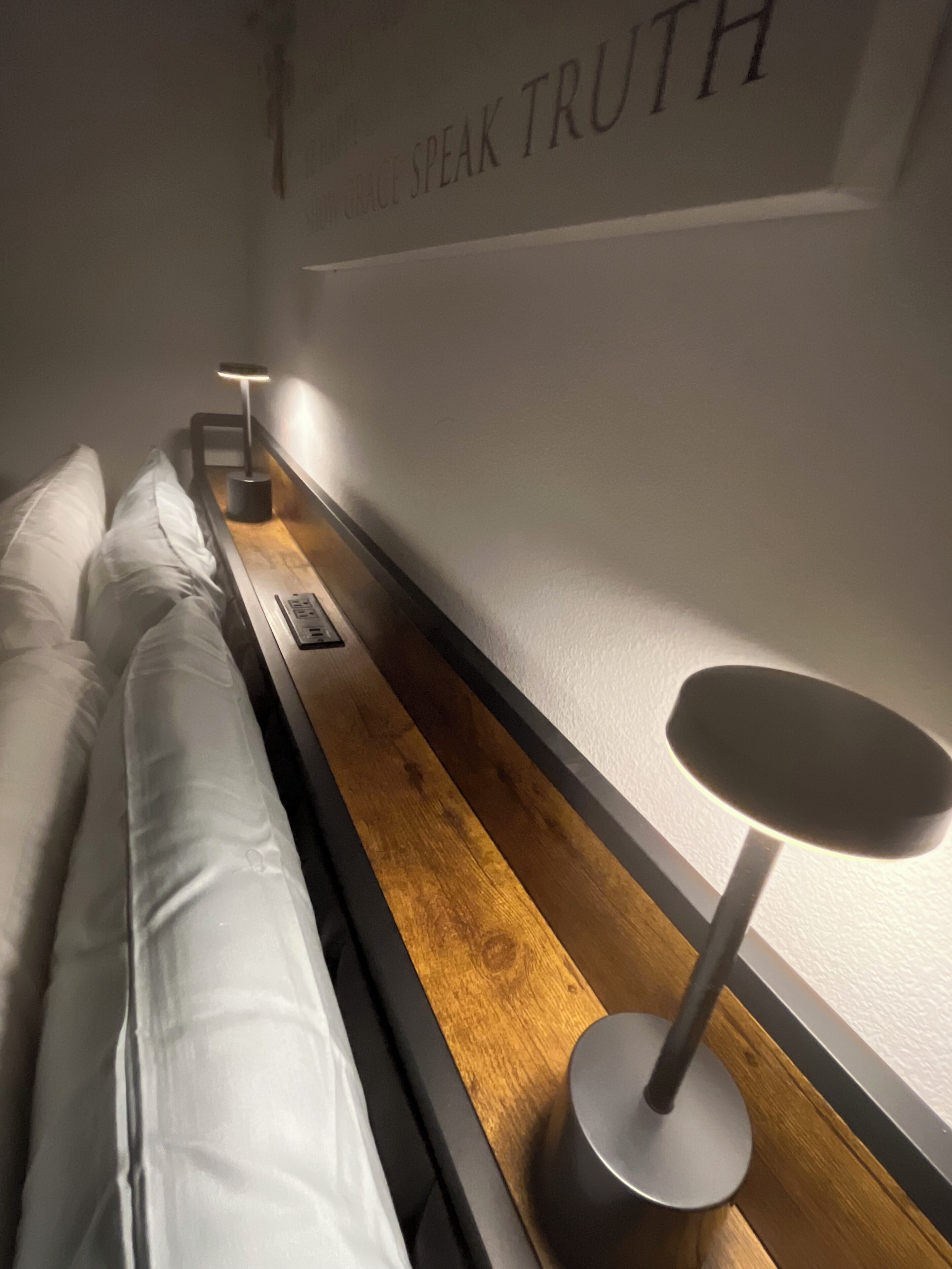 A neatly made bed with white linens, a wooden bedside table, a modern lamp, and a wall with the phrase 'SPEAK TRUTH'.