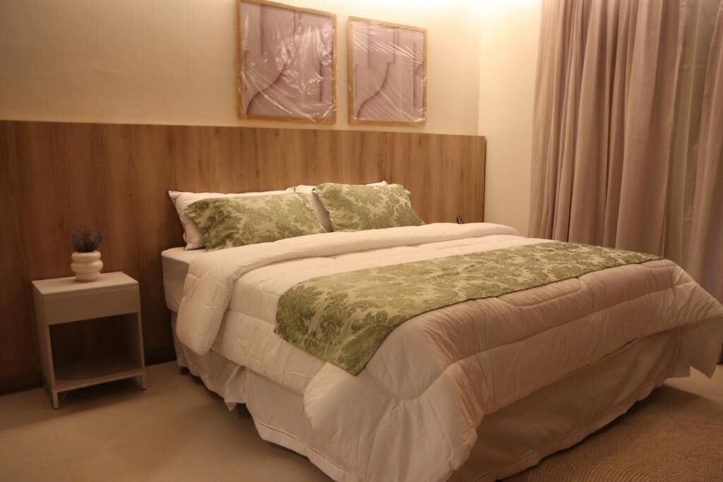 Un lit bien fait, recouvert d’une couverture verte, avec une tête de lit en bois, une petite table de chevet ornée d’un vase et deux tableaux encadrés accrochés au mur.