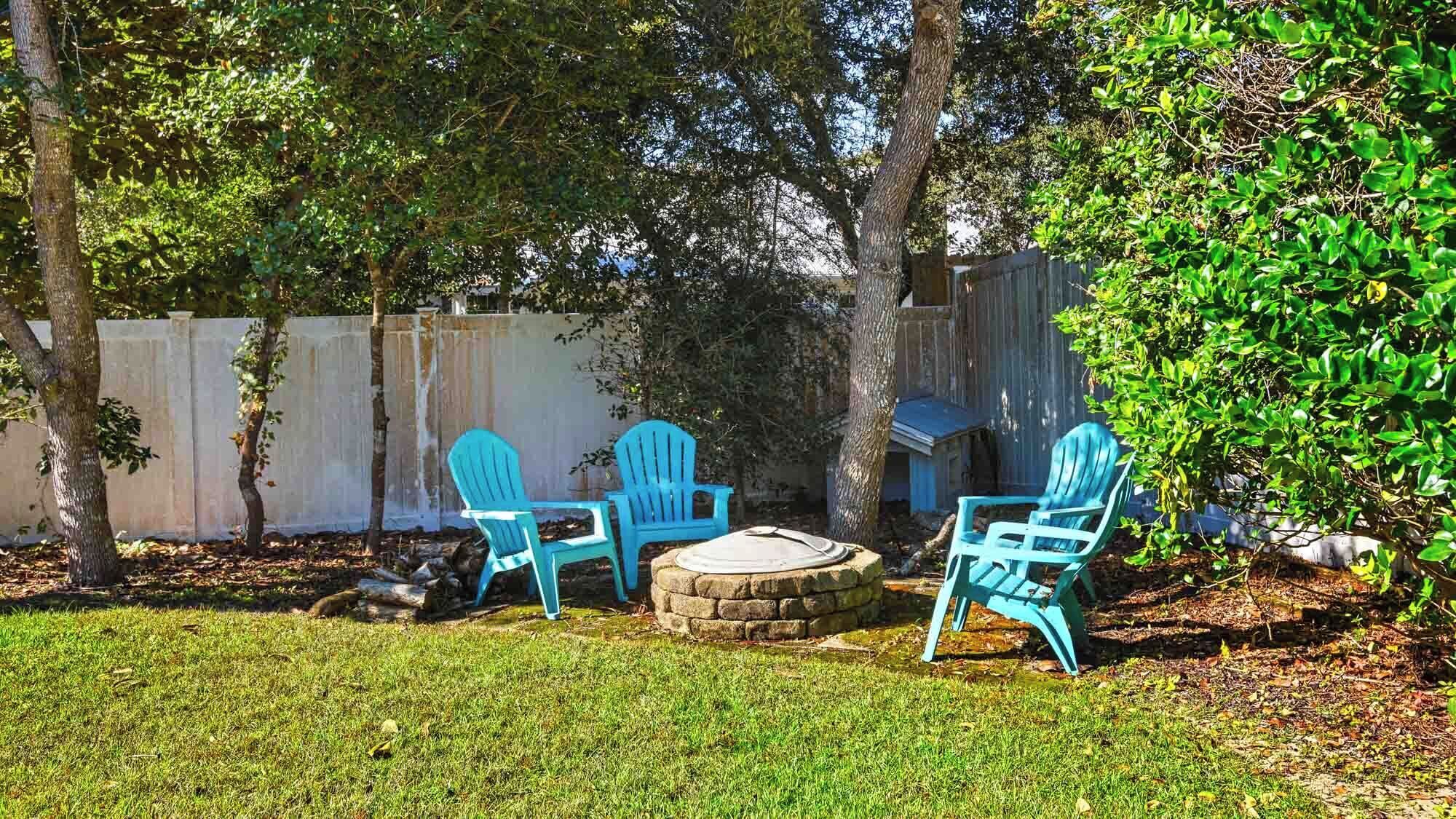 A backyard with a stone fire pit, three blue Adirondack chairs, and a wooden fence.