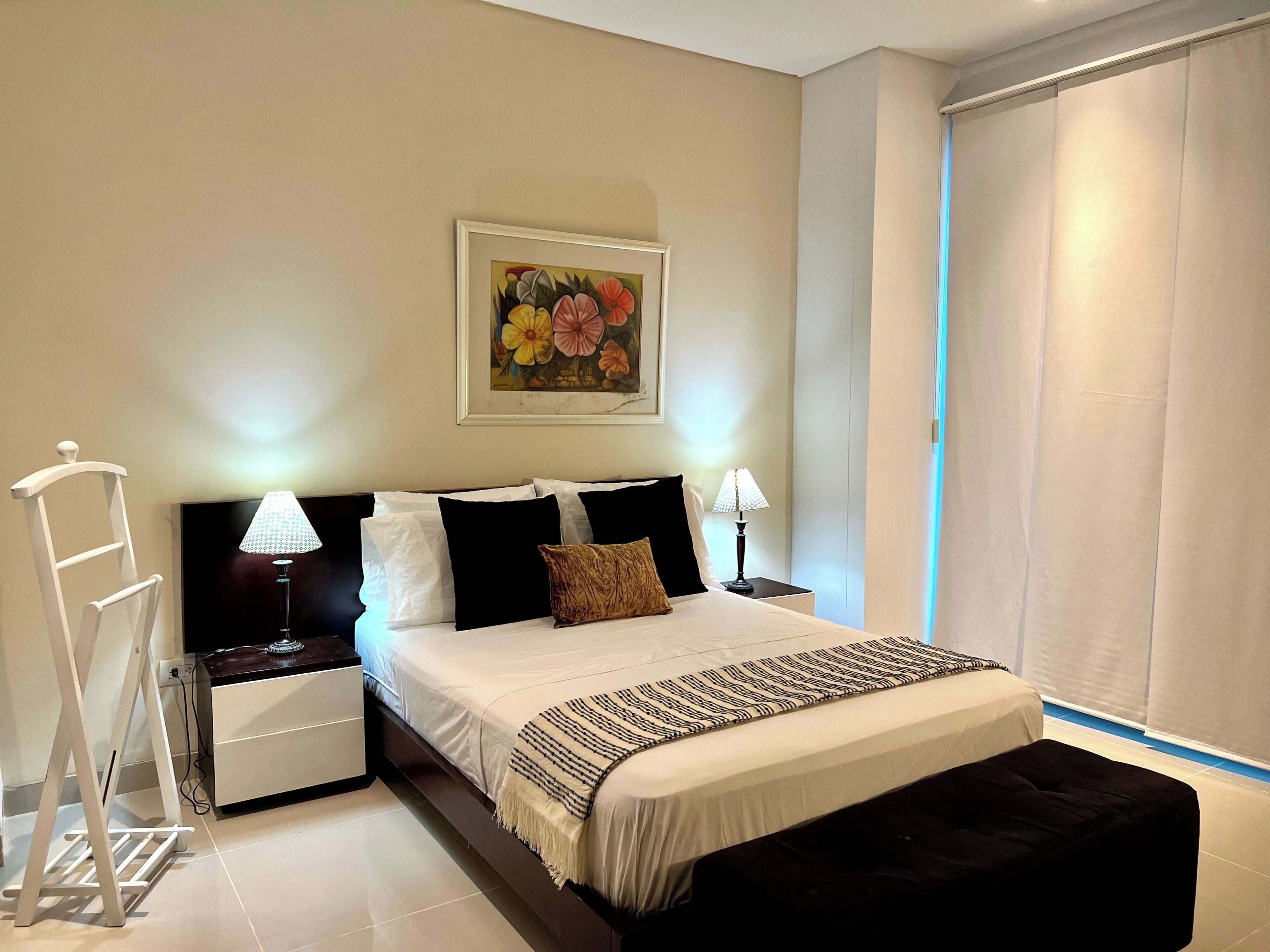 A neatly arranged bedroom with a bed, nightstands, a bedside lamp, a hanging painting, and a window with curtains.