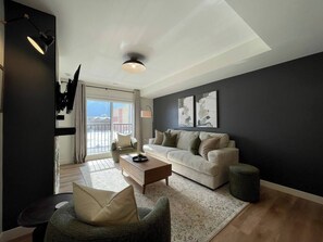 A modern living room with a sofa, armchair, coffee table, and a view of the outdoors through a large window. - Brookside Peaks | Downtown | Rooftop Hot Tub (Canmore)