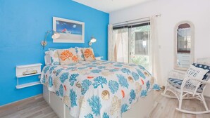 Ein Schlafzimmer mit blauer Wand, einem Bett mit blattgemustertem Überwurf, einem Deckenventilator, einem Fenster mit Vorhängen und einem Korbstuhl mit Kissen.
