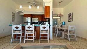 Una cocina con una isla central, armarios de madera, un microondas y un ventilador de techo. Hay cuatro sillas blancas con respaldo estampado alrededor de la isla.
