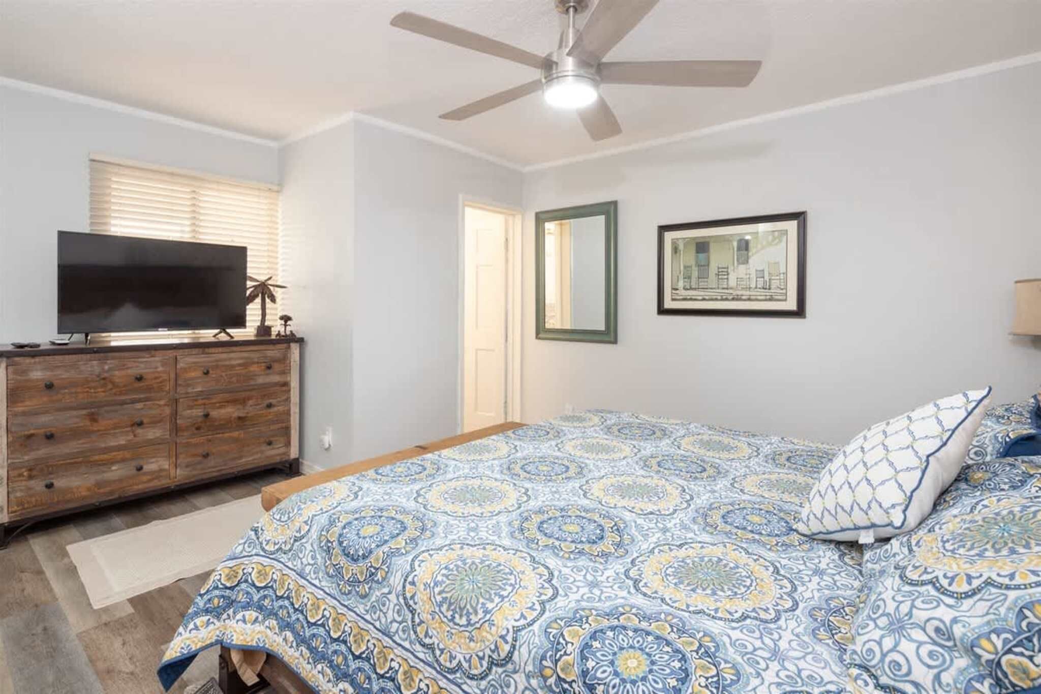 A bedroom with a bed, dresser, television, ceiling fan, and framed artwork.