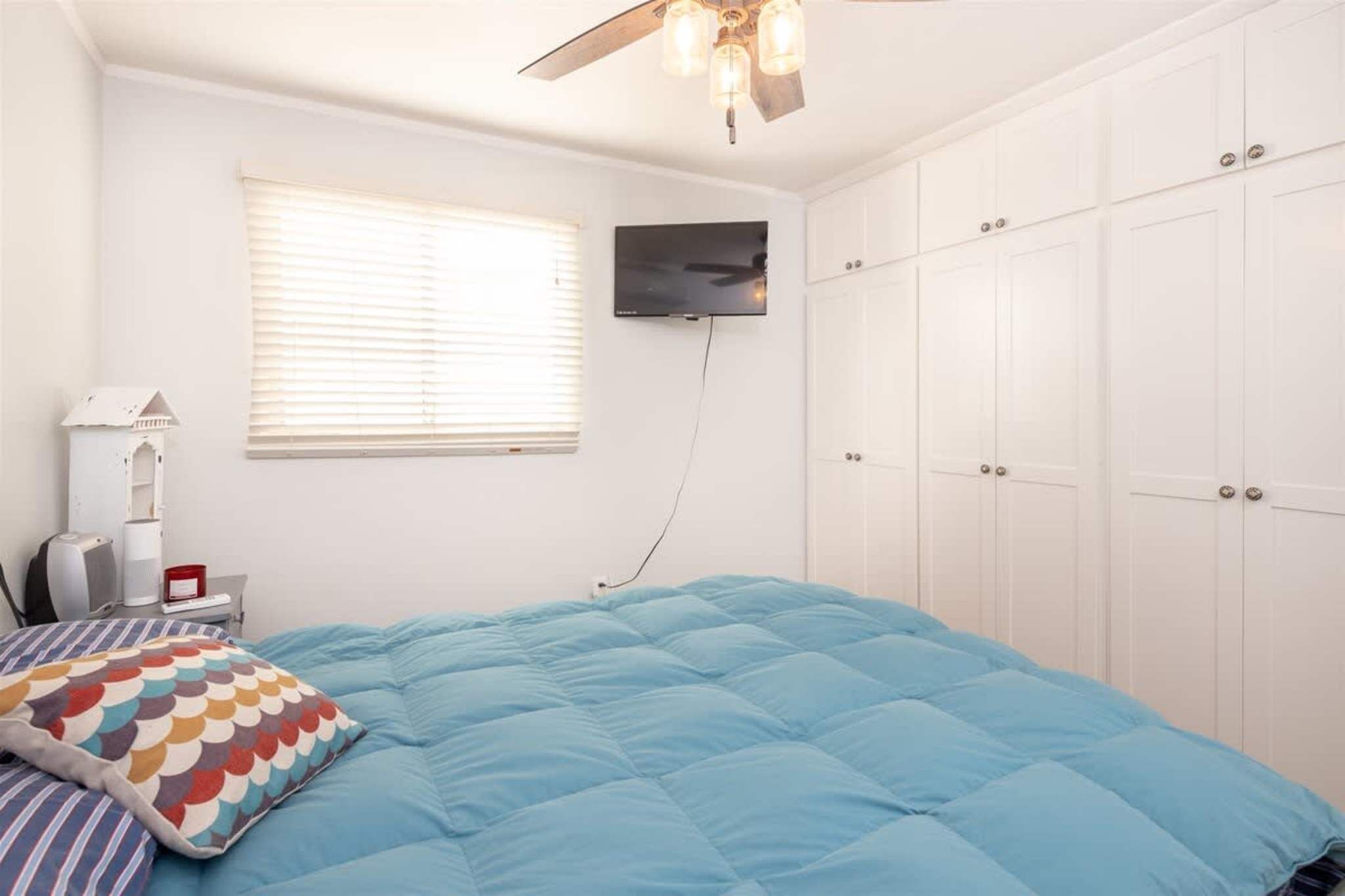 Une chambre à coucher comprenant un lit, un ventilateur de plafond, un téléviseur fixé au mur et une fenêtre avec des stores.