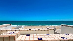 Une vue sur la plage, avec un ciel dégagé, une plage de sable et une série d’espaces de détente recouverts de carreaux blancs et bleus, ornés de motifs bleus.