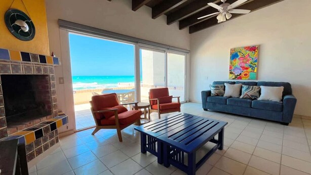 Un salon avec vue sur la plage, une cheminée, un canapé bleu et un tableau coloré.