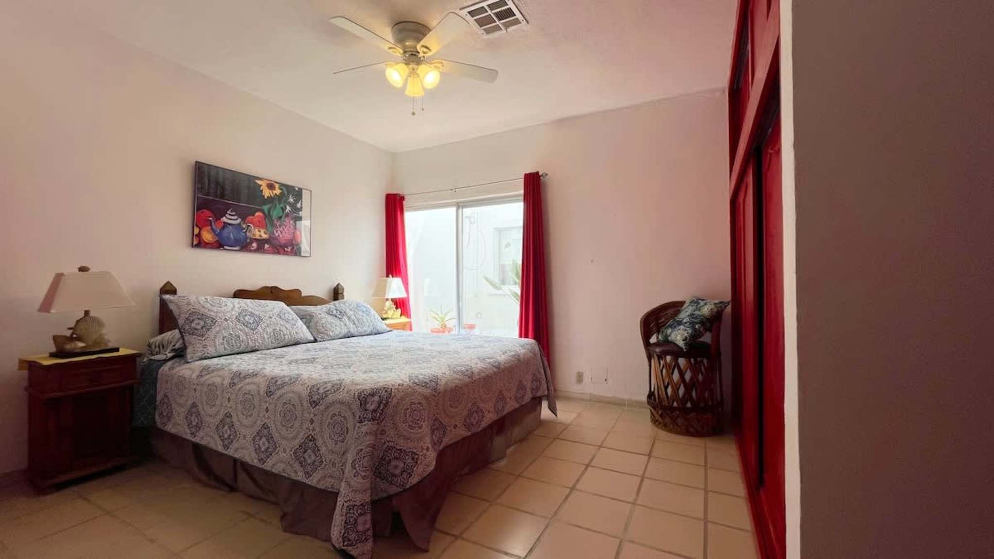 Une chambre à coucher comprenant un lit, une table de chevet, une lampe, un ventilateur de plafond, une fenêtre avec des rideaux et un tableau mural.