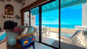 Um quarto com uma cama, um sofá, uma mesinha com desenhos de peixes, uma janela grande com vista para o mar e um ventilador de teto.