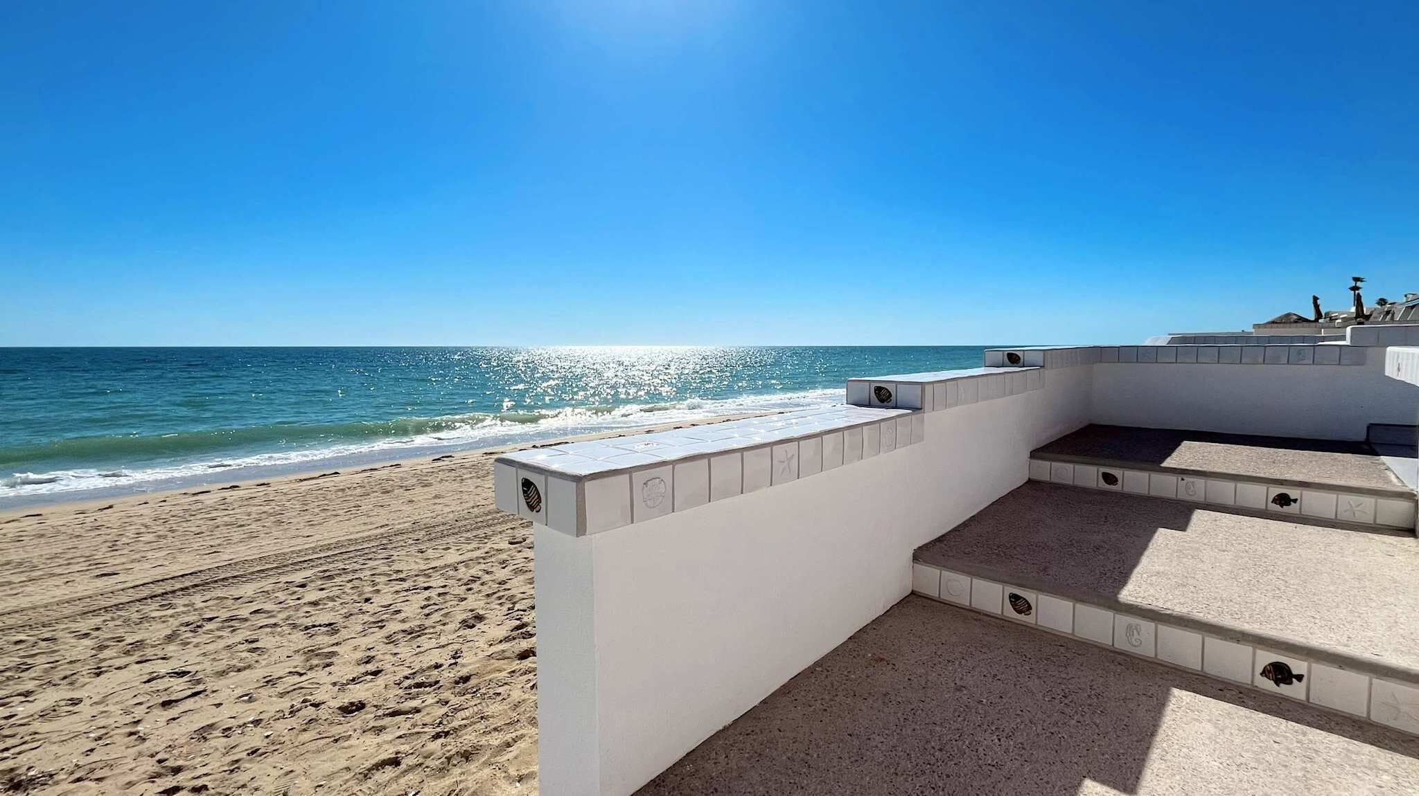 A beachside terrace with a view of the ocean, steps leading down to the sand, and a white wall with decorative tiles.