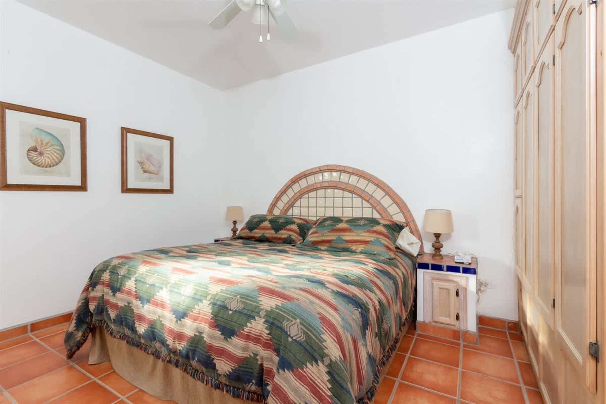 A bedroom with a bed, two framed pictures on the wall, a nightstand with a lamp, and a wooden wardrobe.