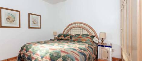 A bedroom with a bed, two framed pictures on the wall, a nightstand with a lamp, and a wooden wardrobe.