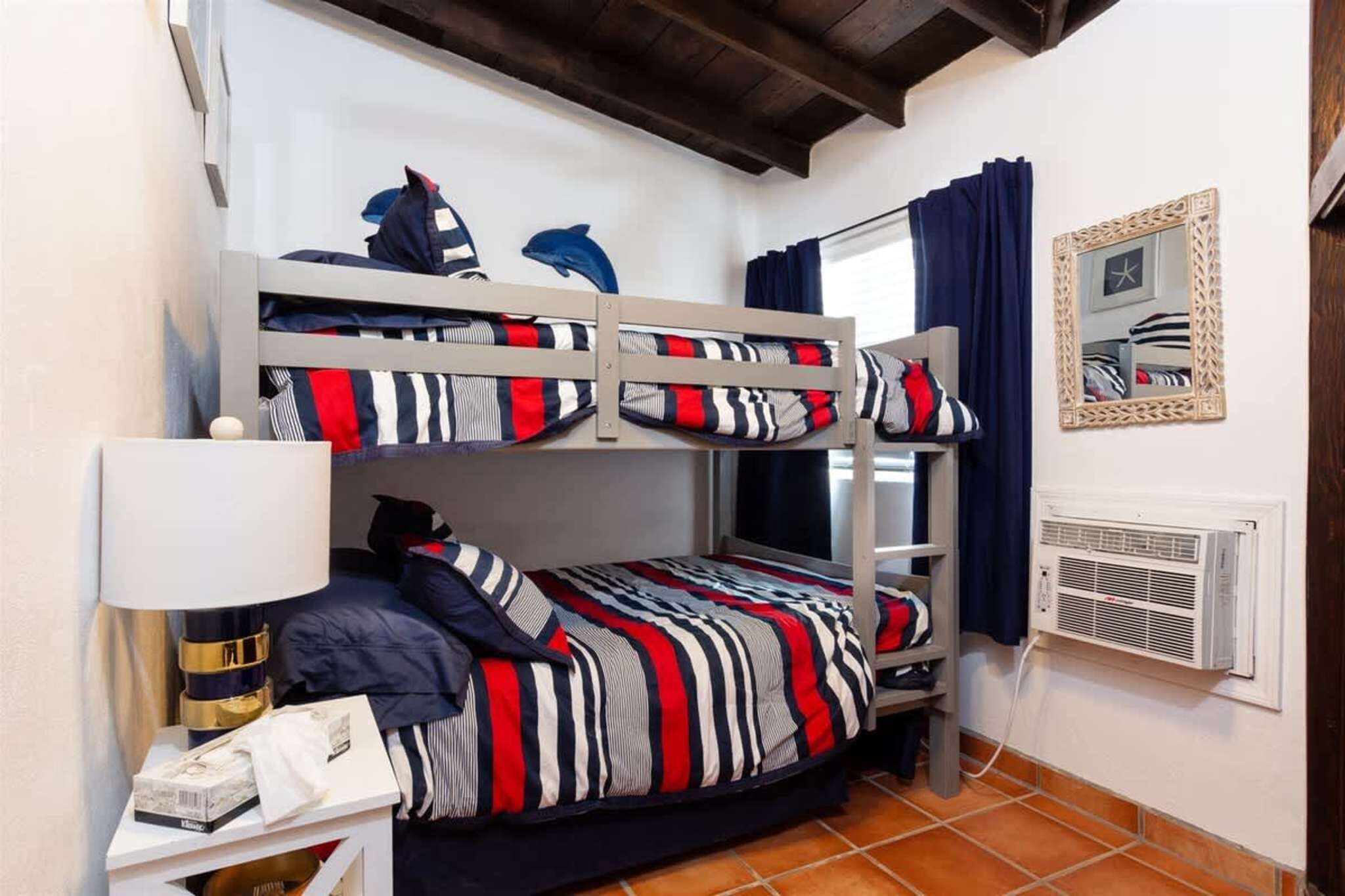 A bunk bed room with striped bedding, a bedside lamp, a mirror, and a window with curtains.