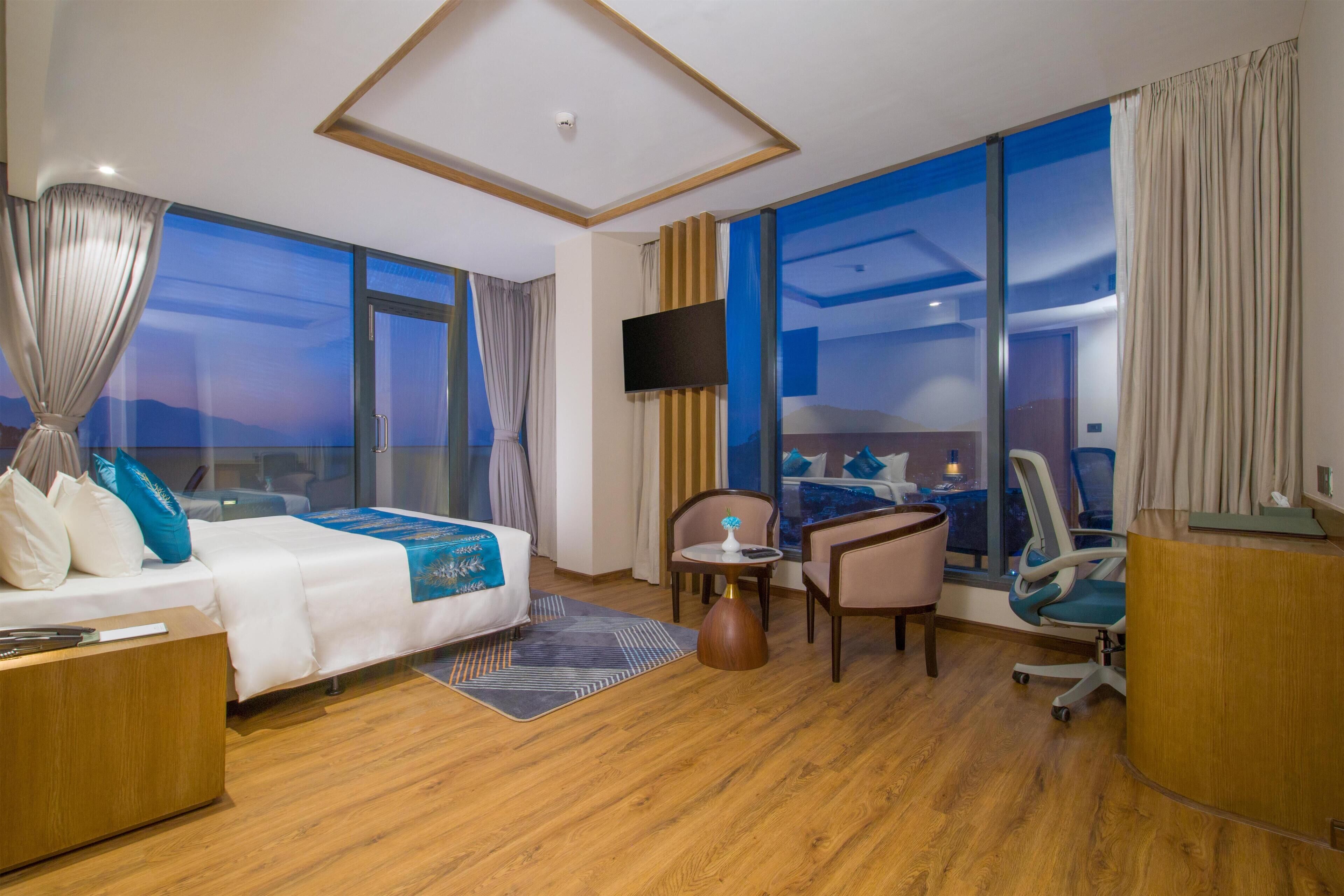 A hotel room with a bed, a desk, a chair, a TV, and large windows.