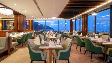 Restaurante moderno com mesas altas e cadeiras estofadas em verde, grandes janelas com vista para a cidade e um candelabro.