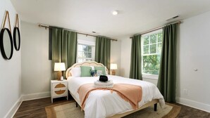 Ein Schlafzimmer mit Bett, Nachttischen, Lampen, einem Fenster mit Vorhängen und einem Wandspiegel.