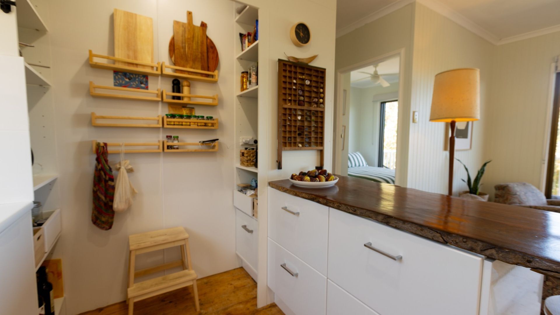 A kitchen with white cabinets, wooden shelves, a small wooden stool, and a bowl of fruit on the counter.