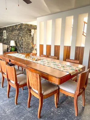 A dining area with a long wooden table, cushioned chairs, a stone wall, and a window with blinds. - Private Group Retreat • 350m to Olas Altas-Balcony & Stunning Views (Mazatlán)