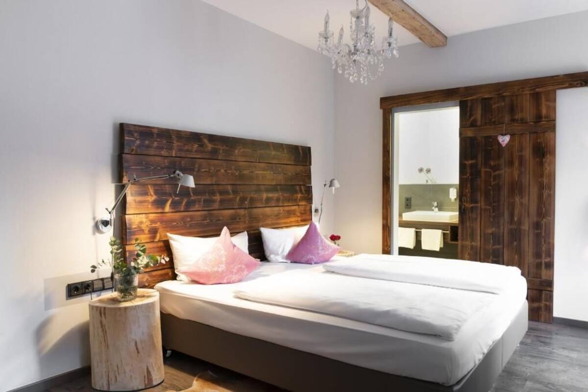 A bedroom with a wooden headboard, a bedside table with a lamp, a large bed with pillows, a wooden floor, and a chandelier.