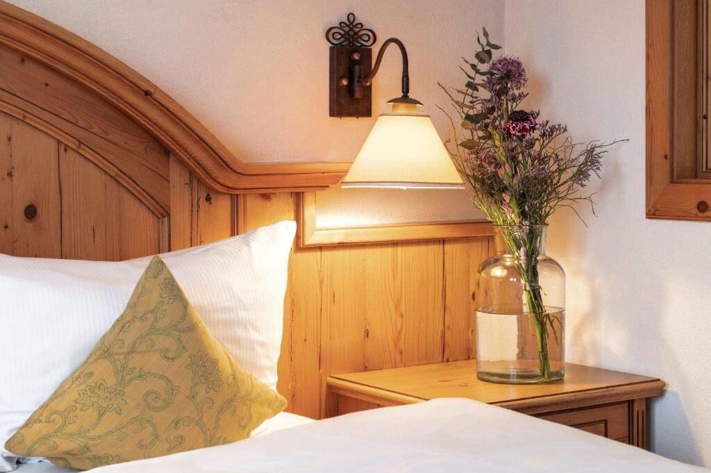 Una habitación de hotel con cabecera de madera, lámpara de noche, un florero con flores y una mesita de noche.