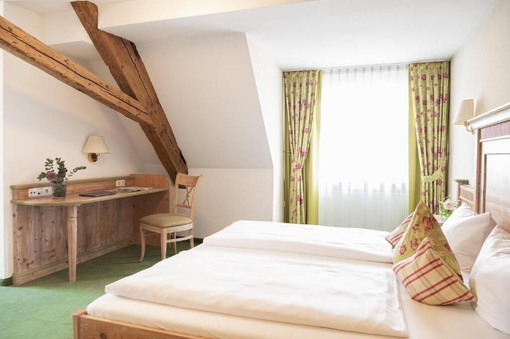 Ein Schlafzimmer mit einem Holztisch, einem Stuhl, einem Bett mit weißer Bettwäsche und einem Fenster mit blumigen Vorhängen.