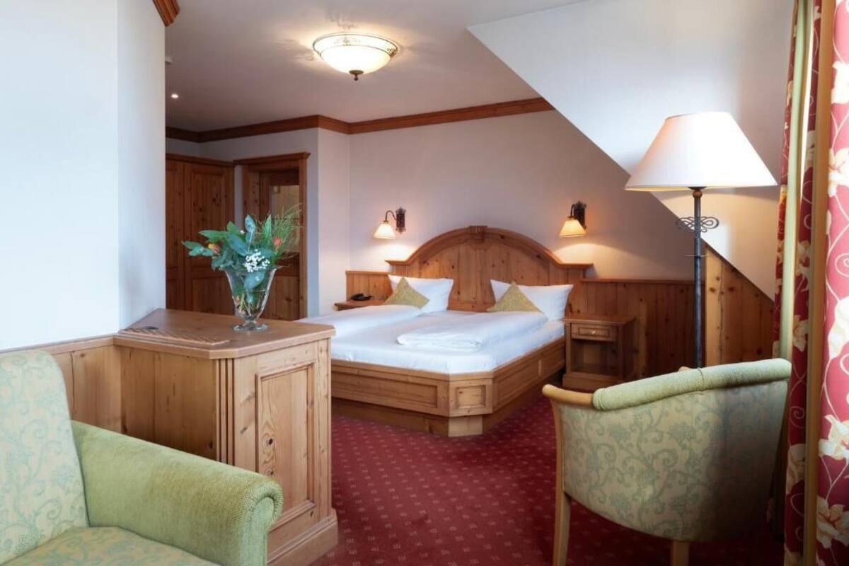 A bedroom with a wooden bed, a nightstand, a lamp, a chair, and a vase with flowers.