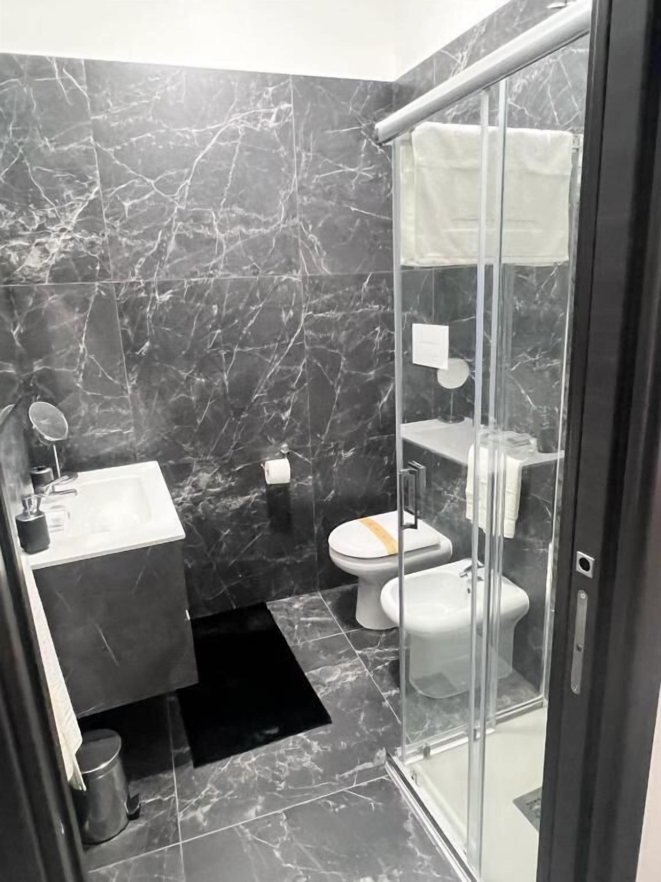 Un baño moderno con cabina de ducha de vidrio, inodoro blanco, lavamanos con espejo y paredes de mármol.