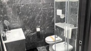 Un baño moderno con cabina de ducha de vidrio, inodoro blanco, lavamanos con espejo y paredes de mármol.