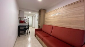 A modern living room with a red sofa, a wooden accent wall, and a kitchen area in the background. - Aitzina Studio - Downtown (CAMBO LES BAINS)