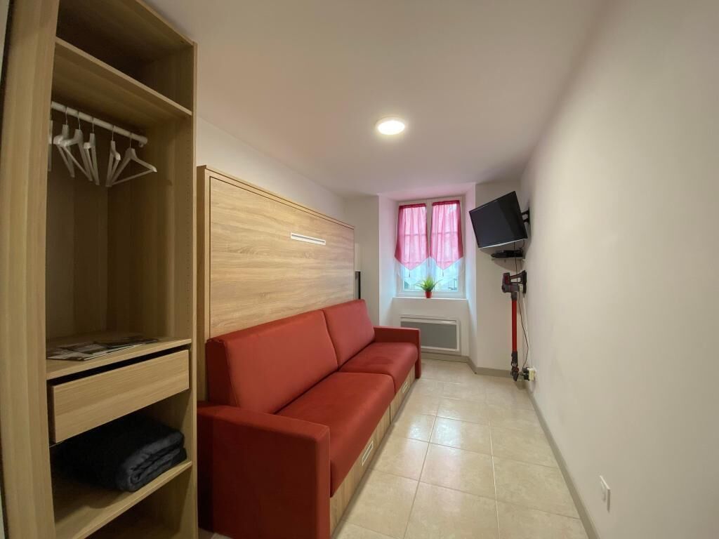 Une pièce avec une armoire en bois, un canapé rouge, un téléviseur fixé au mur et une fenêtre ornée de rideaux roses.
