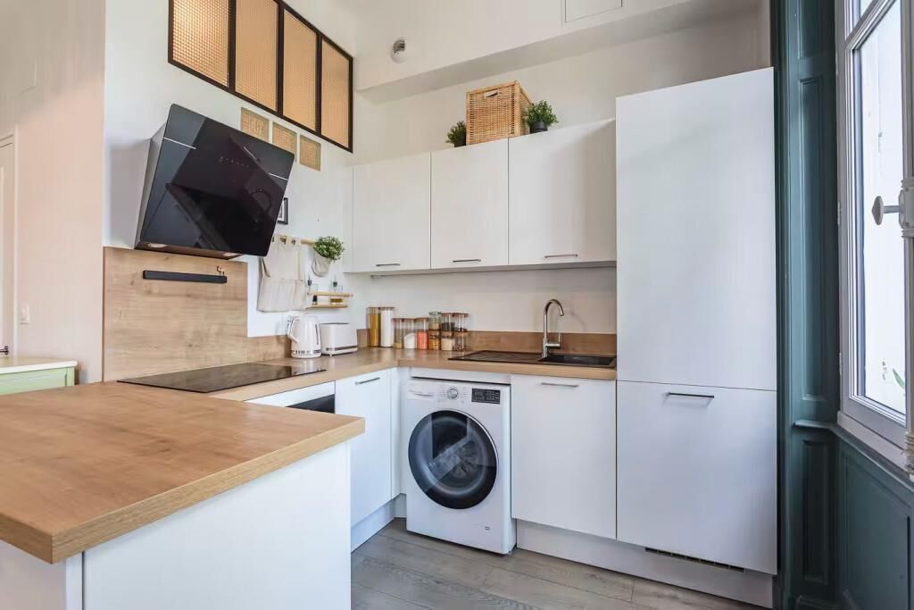 Eine kompakte Küche mit Waschmaschine, weißen Schränken, einer Holz Arbeitsplatte und einem großen Fenster.