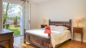 A bedroom with a bed, two bedside tables, a wooden dresser, and a sliding glass door leading to a garden.