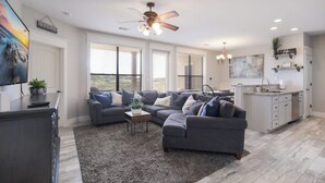 Ein modernes Wohnzimmer mit einem grauen Ecksofa, einem Deckenventilator und einer Küchenzeile mit weißen Schränken und einer Arbeitsplatte.